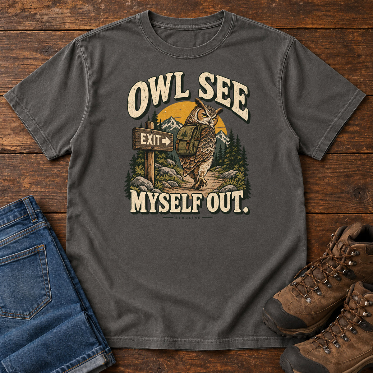 Pepper Owl See Myself Out T-Shirt with an owl wearing a backpack beside an Exit sign on a woodland trail; front flat lay on wood.