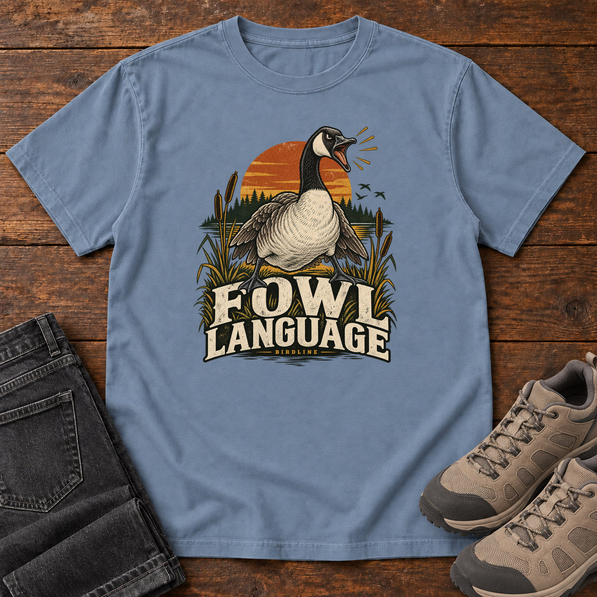 Blue Jean Fowl Language T-Shirt with an open-beaked goose among reeds against an orange sunset; front flat lay on wood.