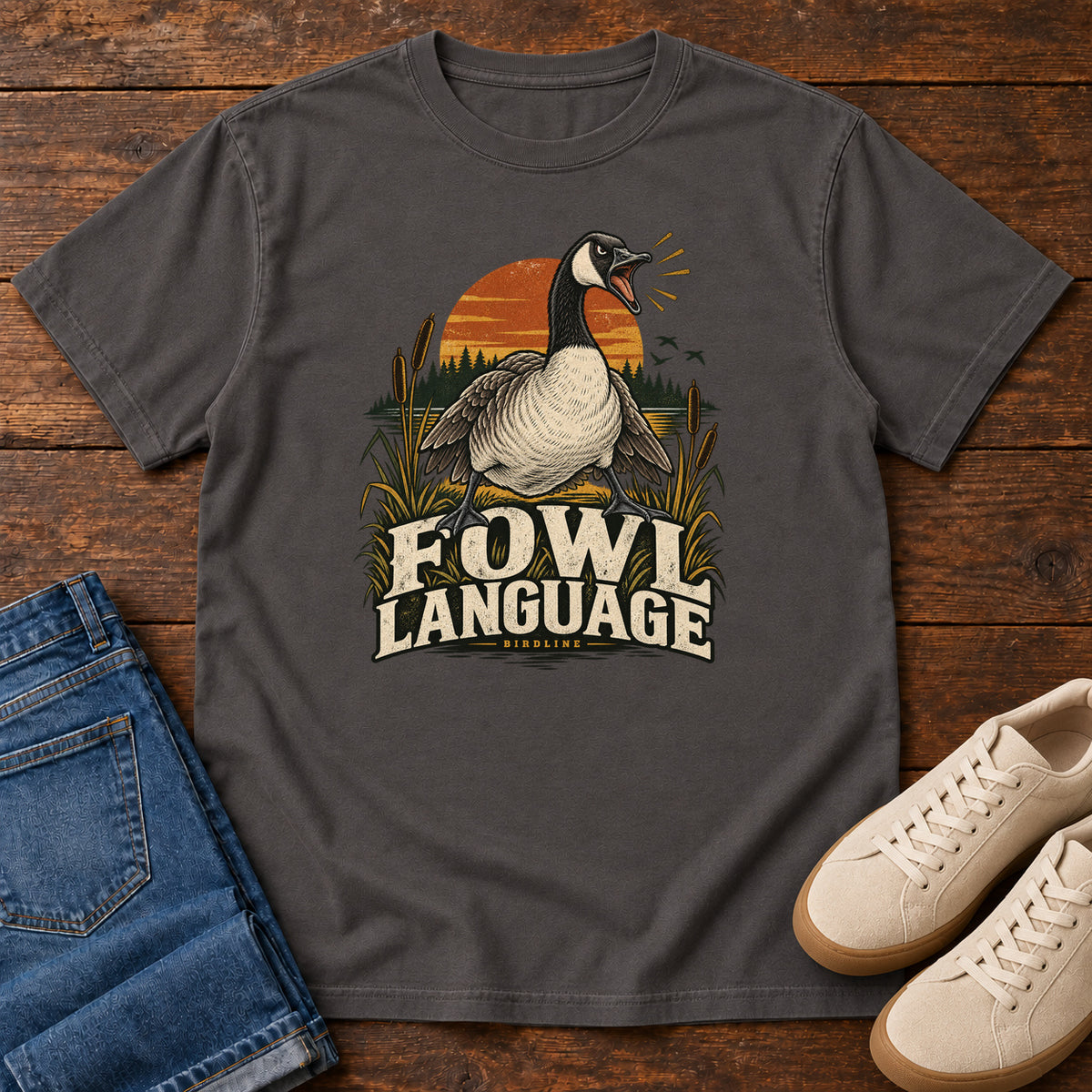 Graphite Fowl Language T-Shirt with an open-beaked goose among reeds against an orange sunset; front flat lay on wood.