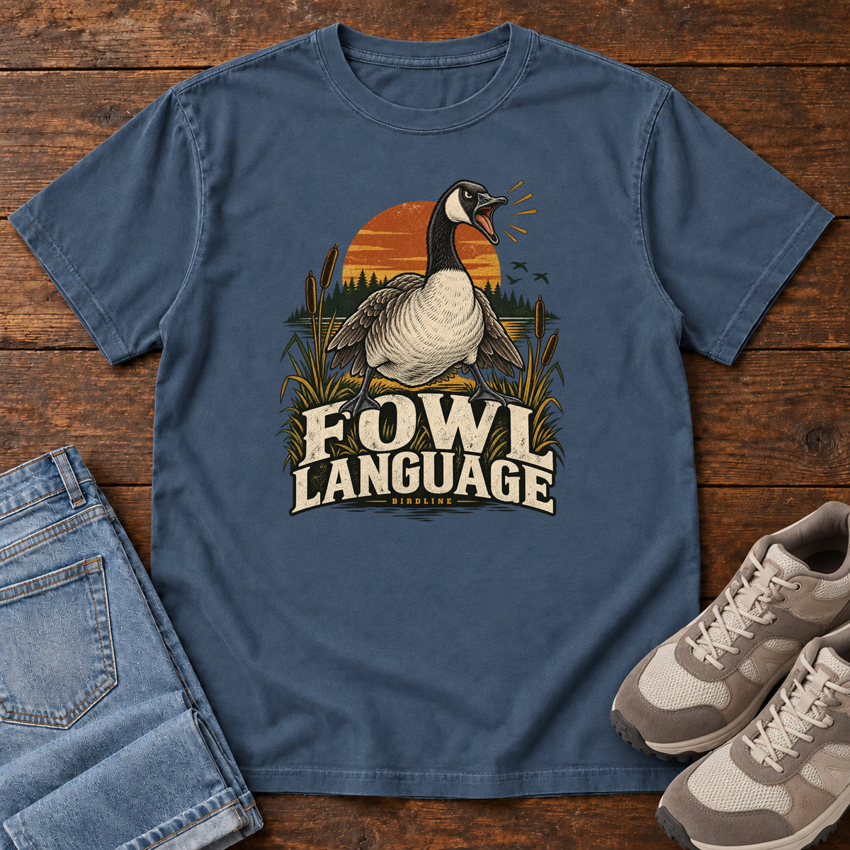 Midnight Fowl Language T-Shirt with an open-beaked goose among reeds against an orange sunset; front flat lay on wood.