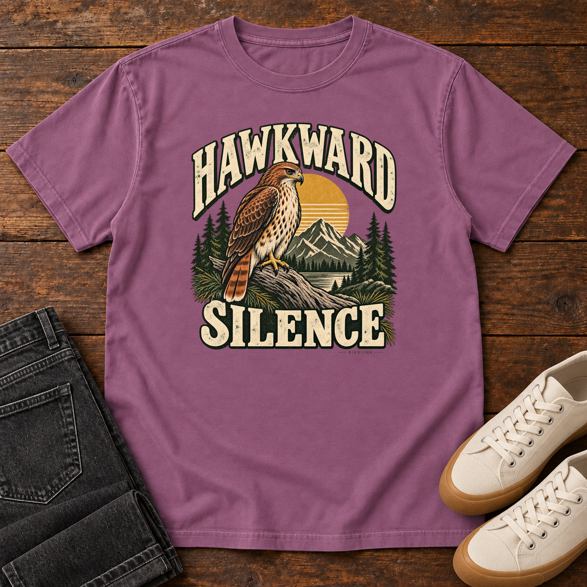 Berry Hawkward Silence T-Shirt with a brown bird of prey perched above a mountain lake between cream Hawkward Silence lettering, front flat lay on wood.