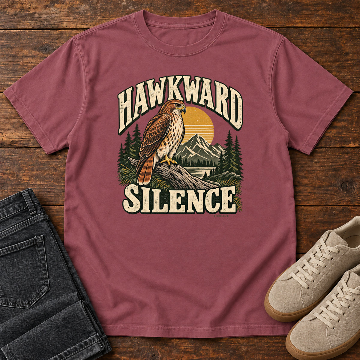Brick Hawkward Silence T-Shirt with a brown bird of prey perched above a mountain lake between cream Hawkward Silence lettering, front flat lay on wood.