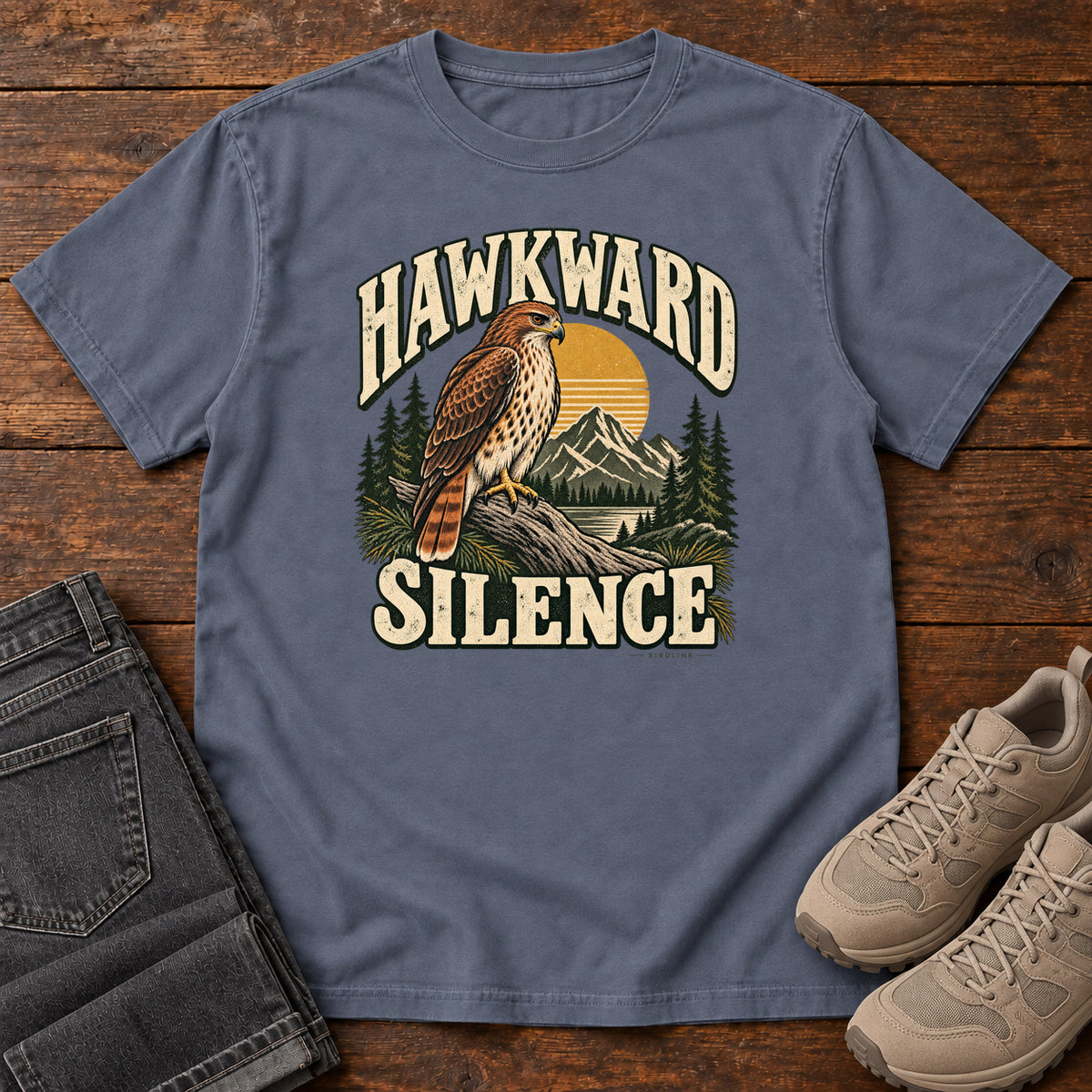 Denim Hawkward Silence T-Shirt with a brown bird of prey perched above a mountain lake between cream Hawkward Silence lettering, front flat lay on wood.