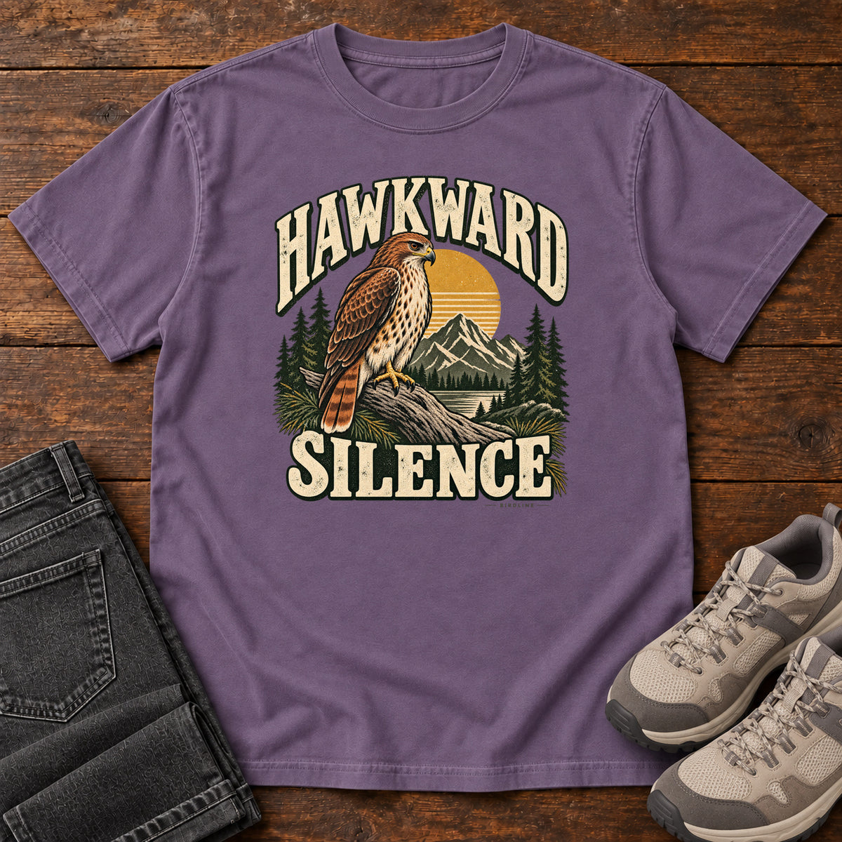 Grape Hawkward Silence T-Shirt with a brown bird of prey perched above a mountain lake between cream Hawkward Silence lettering, front flat lay on wood.