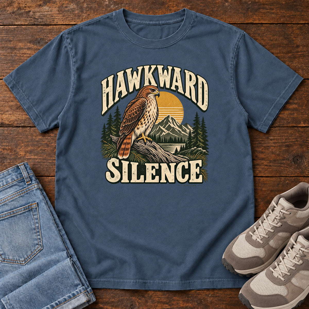 Midnight Hawkward Silence T-Shirt with a brown bird of prey perched above a mountain lake between cream Hawkward Silence lettering, front flat lay on wood.