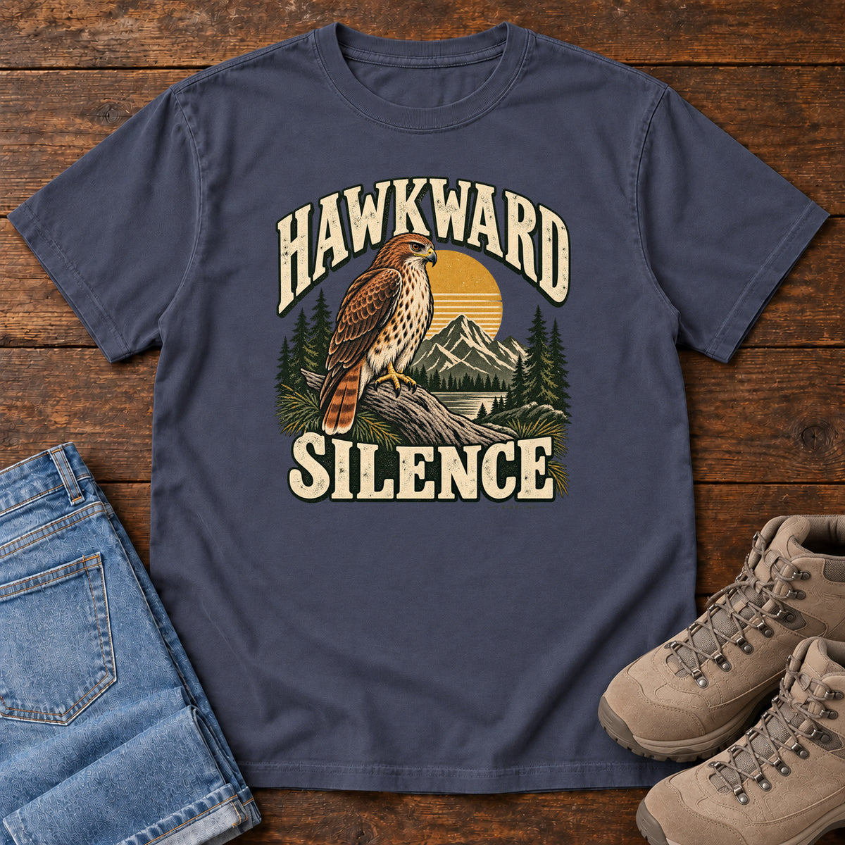 Navy Hawkward Silence T-Shirt with a brown bird of prey perched above a mountain lake between cream Hawkward Silence lettering, front flat lay on wood.