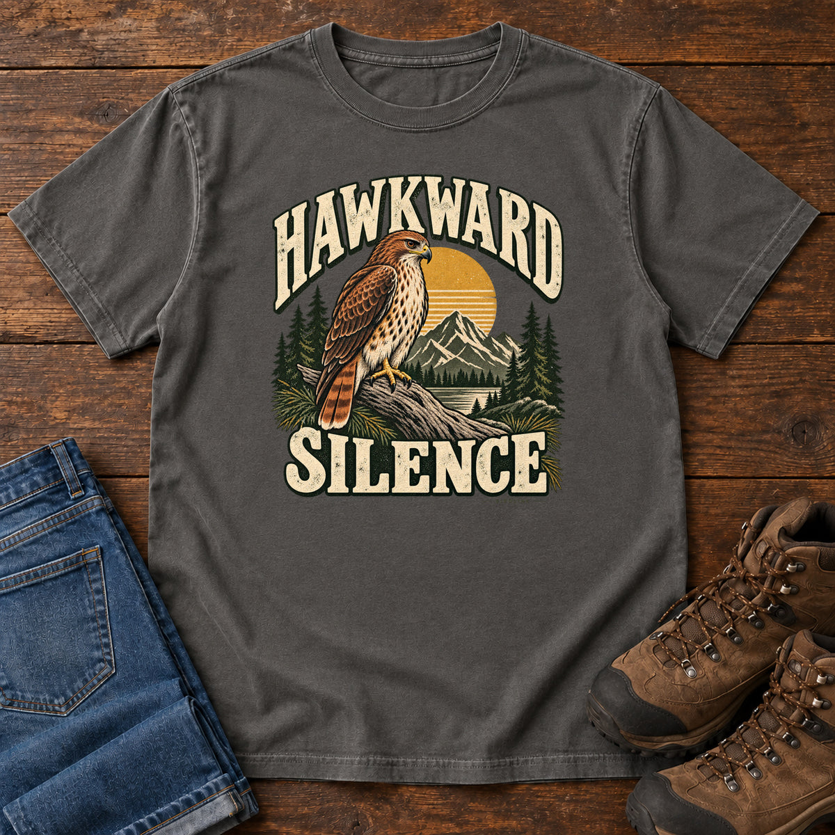 Pepper Hawkward Silence T-Shirt with a brown bird of prey perched above a mountain lake between cream Hawkward Silence lettering, front flat lay on wood.