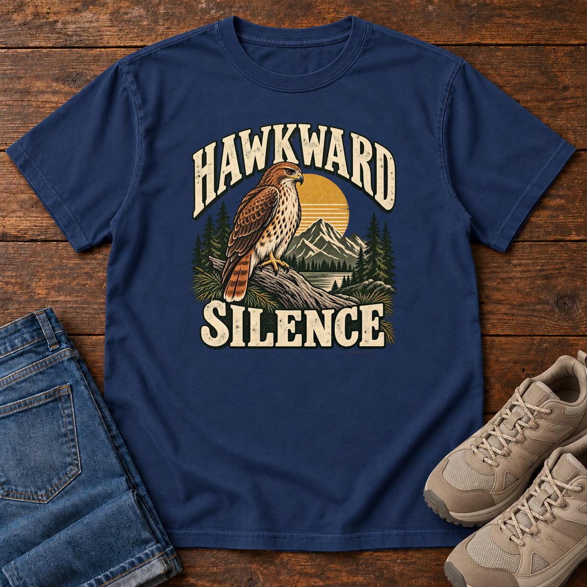 True Navy Hawkward Silence T-Shirt with a brown bird of prey perched above a mountain lake between cream Hawkward Silence lettering, front flat lay on wood.