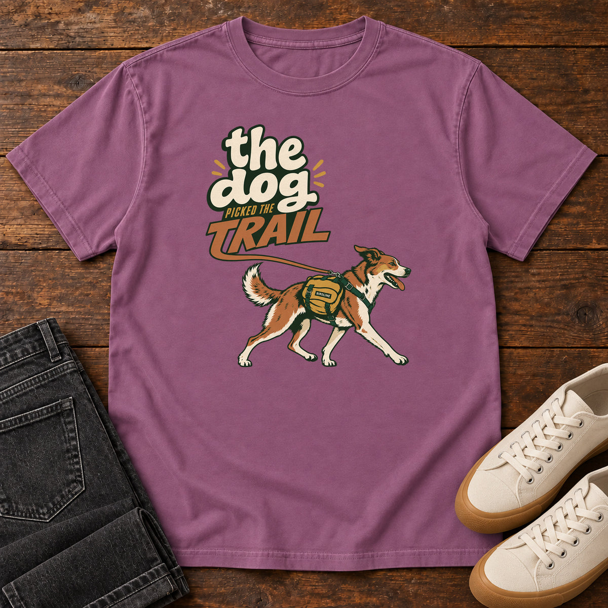 The Dog Picked the Trail T-Shirt in Berry, front laid flat on wood, featuring a brown-and-white dog walking with a hiking pack beneath cream-and-orange lettering.