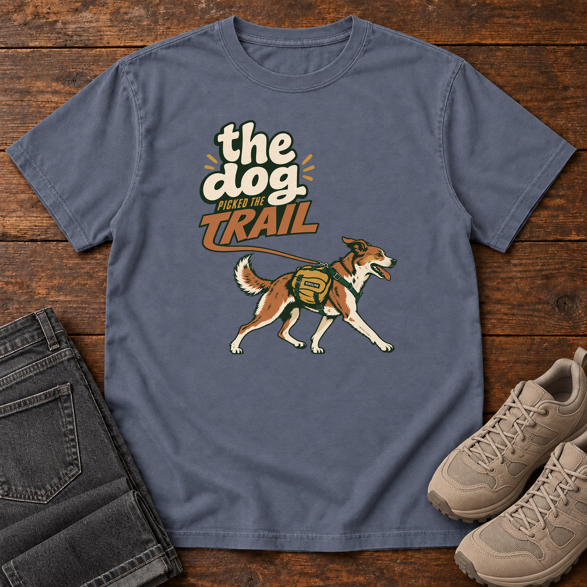 The Dog Picked the Trail T-Shirt in Denim, front laid flat on wood, featuring a brown-and-white dog walking with a hiking pack beneath cream-and-orange lettering.