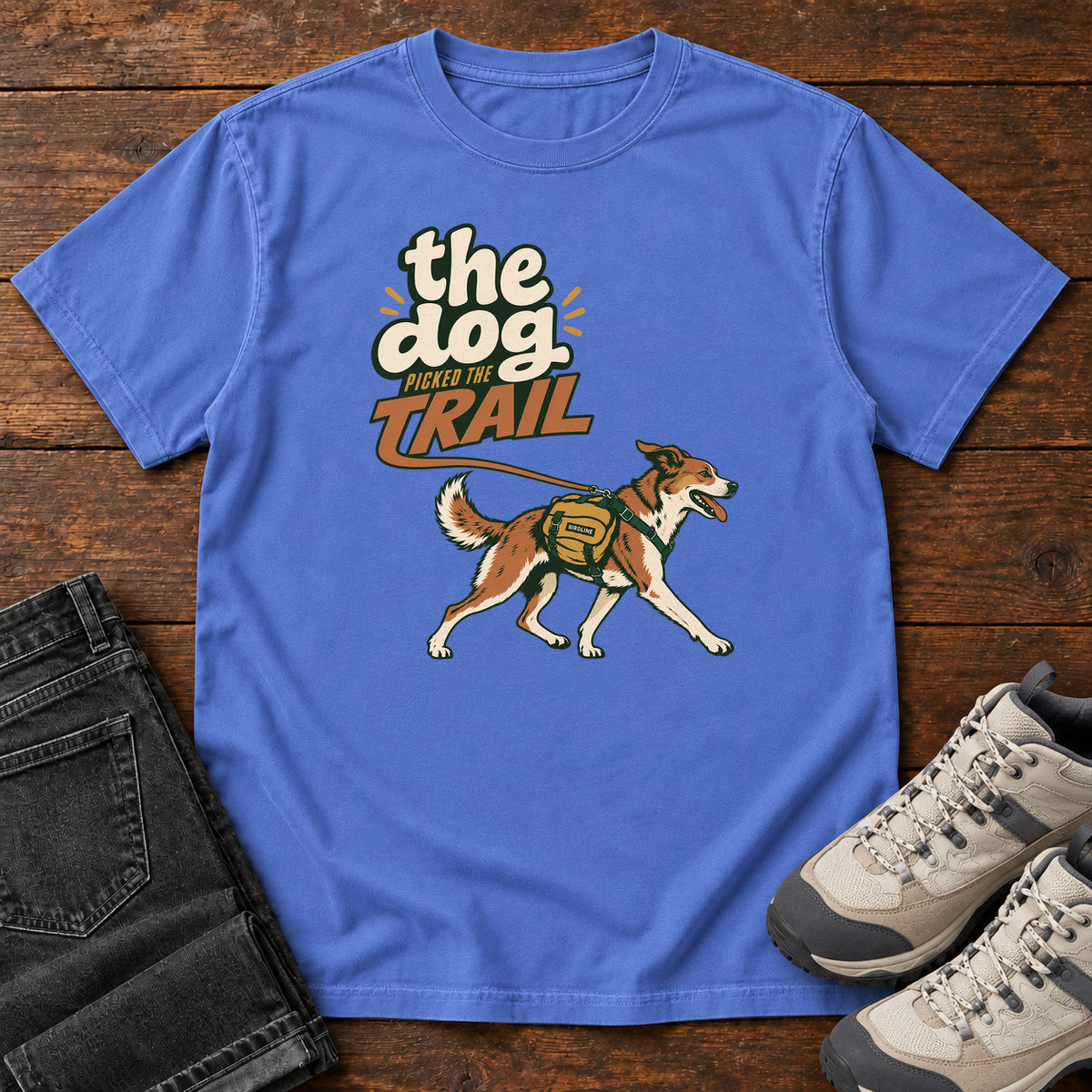The Dog Picked the Trail T-Shirt in Mystic Blue, front laid flat on wood, featuring a brown-and-white dog walking with a hiking pack beneath cream-and-orange lettering.