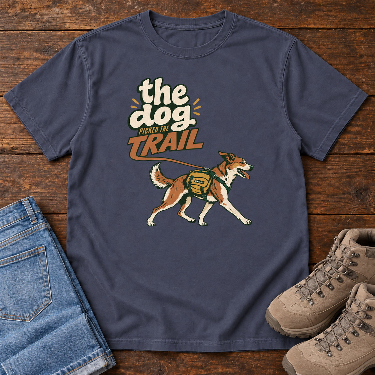 The Dog Picked the Trail T-Shirt in Navy, front laid flat on wood, featuring a brown-and-white dog walking with a hiking pack beneath cream-and-orange lettering.