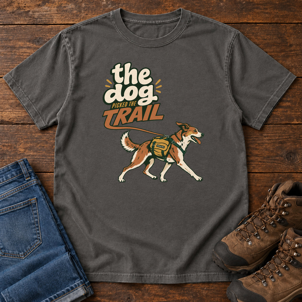The Dog Picked the Trail T-Shirt in Pepper, front laid flat on wood, featuring a brown-and-white dog walking with a hiking pack beneath cream-and-orange lettering.