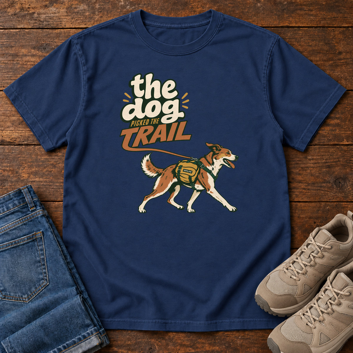 The Dog Picked the Trail T-Shirt in True Navy, front laid flat on wood, featuring a brown-and-white dog walking with a hiking pack beneath cream-and-orange lettering.