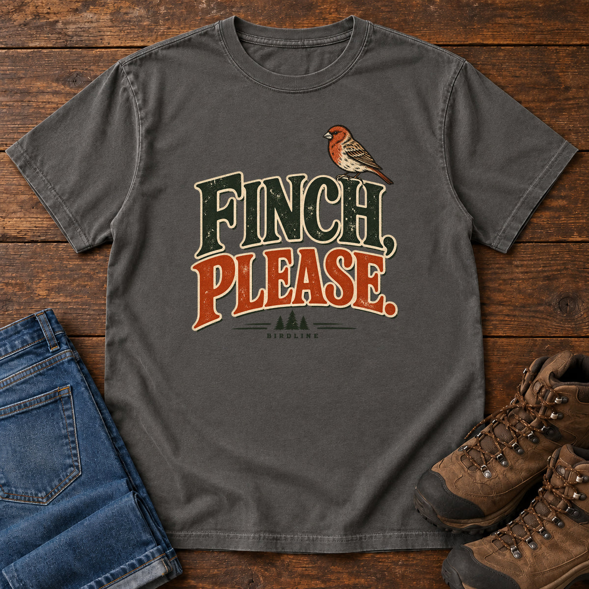 Pepper Finch, Please. T-Shirt with a small bird perched above cream-outlined green-and-orange lettering, front flat lay on wood.
