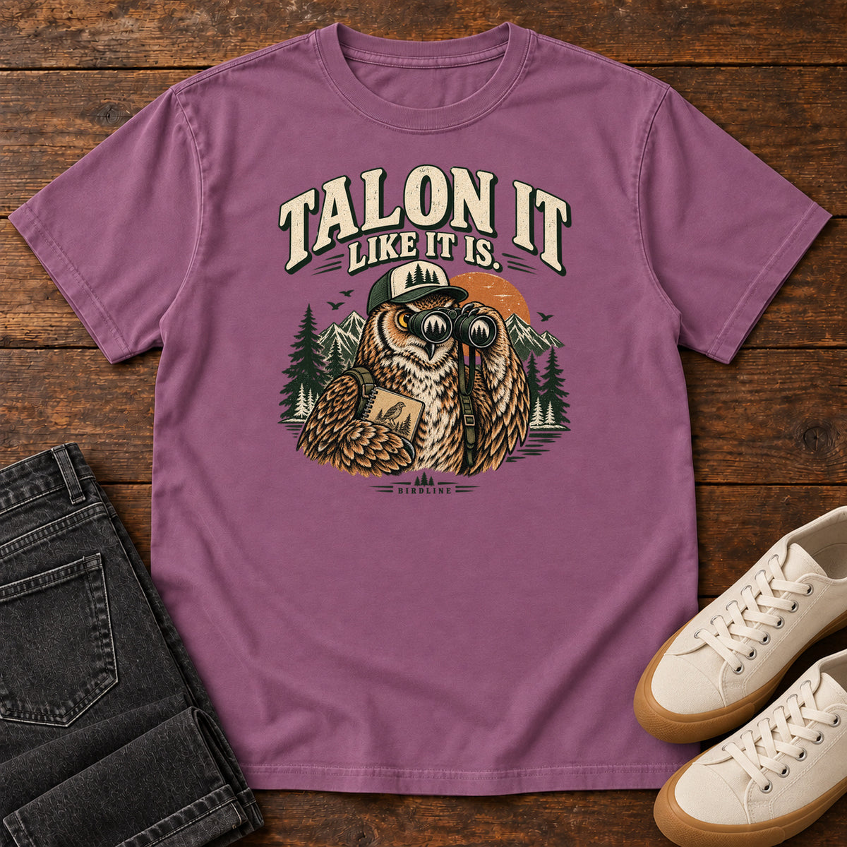 Berry Talon It Like It Is. T-Shirt with an owl wearing a cap, looking through binoculars and holding a field notebook; front flat lay on wood.