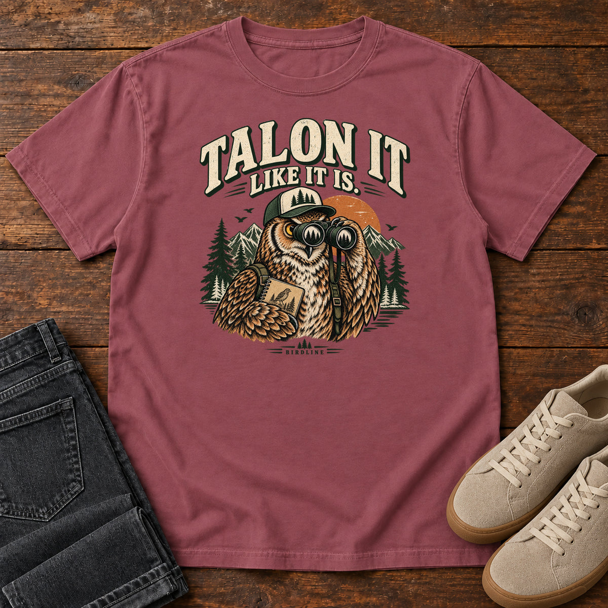 Brick Talon It Like It Is. T-Shirt with an owl wearing a cap, looking through binoculars and holding a field notebook; front flat lay on wood.