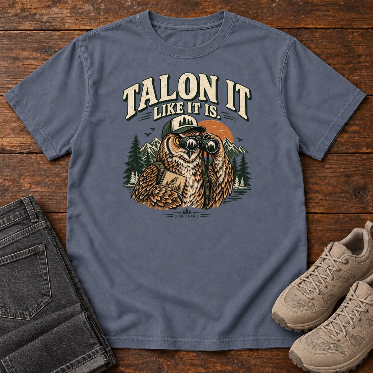 Denim Talon It Like It Is. T-Shirt with an owl wearing a cap, looking through binoculars and holding a field notebook; front flat lay on wood.