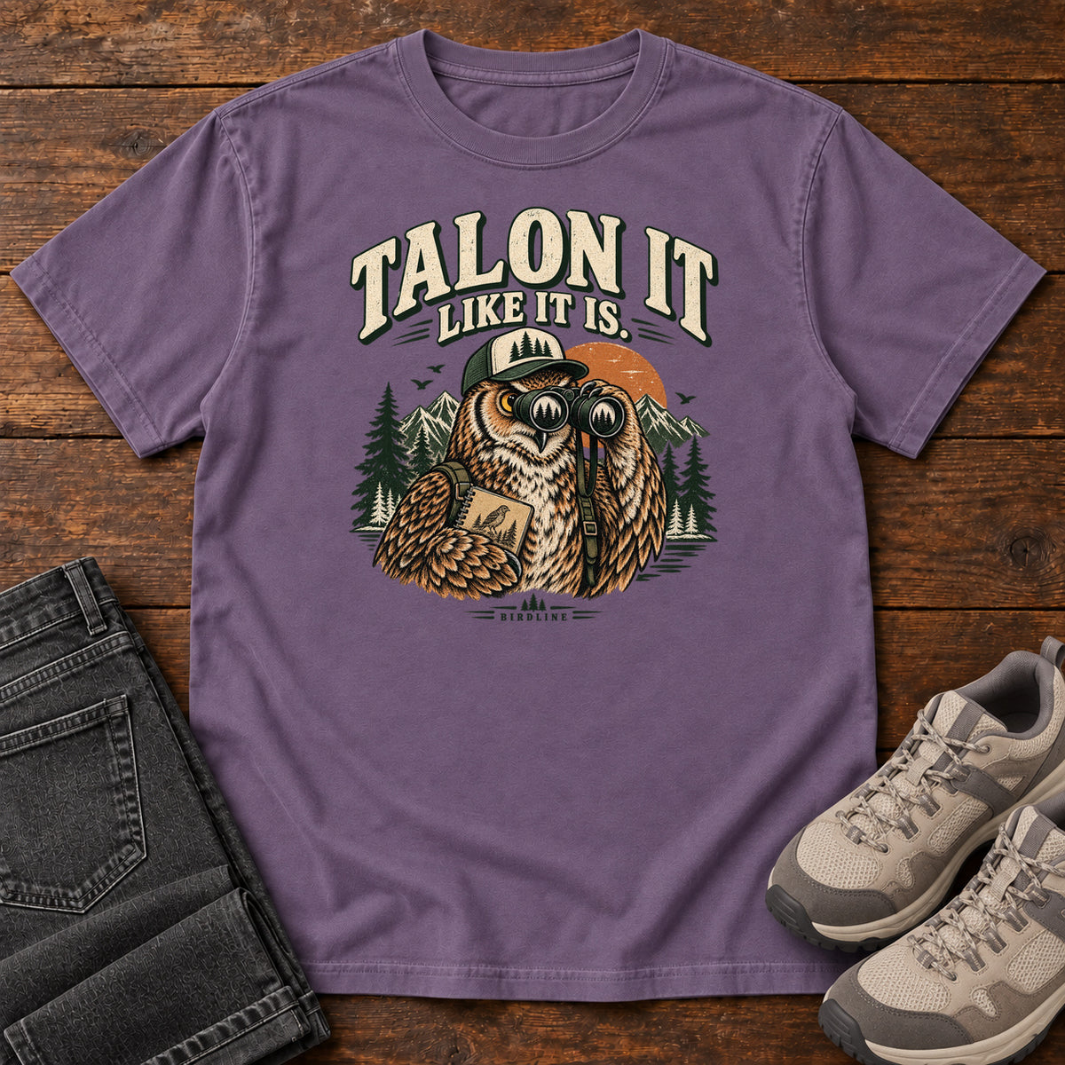 Grape Talon It Like It Is. T-Shirt with an owl wearing a cap, looking through binoculars and holding a field notebook; front flat lay on wood.
