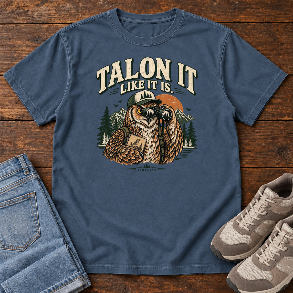 Midnight Talon It Like It Is. T-Shirt with an owl wearing a cap, looking through binoculars and holding a field notebook; front flat lay on wood.