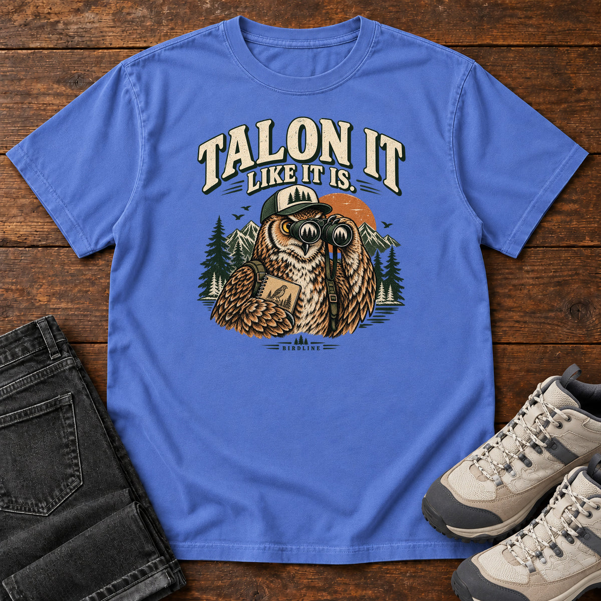 Mystic Blue Talon It Like It Is. T-Shirt with an owl wearing a cap, looking through binoculars and holding a field notebook; front flat lay on wood.