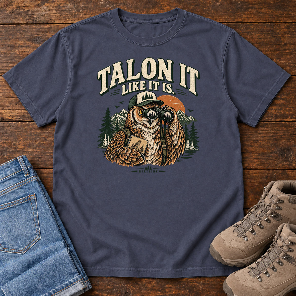 Navy Talon It Like It Is. T-Shirt with an owl wearing a cap, looking through binoculars and holding a field notebook; front flat lay on wood.
