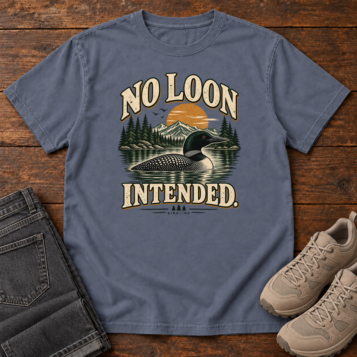 Denim No Loon Intended T-Shirt with a black-and-white waterbird on a mountain lake beneath an orange sun and cream lettering, front flat lay on wood.