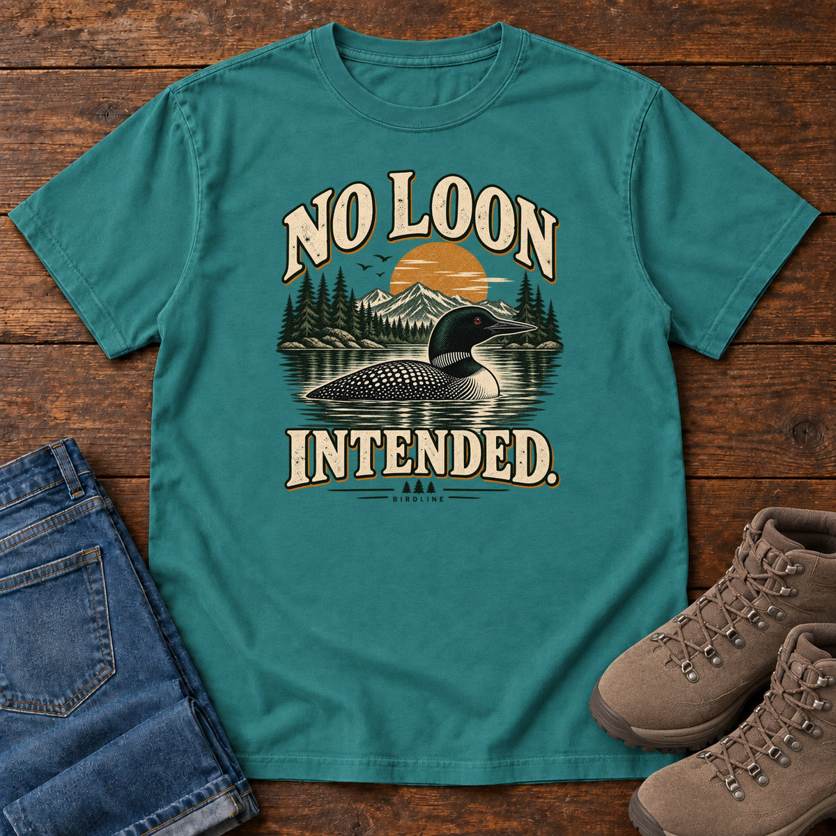 Emerald No Loon Intended T-Shirt with a black-and-white waterbird on a mountain lake beneath an orange sun and cream lettering, front flat lay on wood.