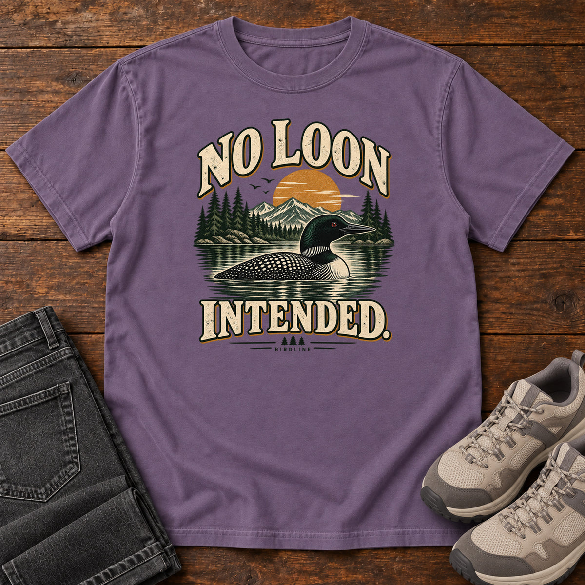 Grape No Loon Intended T-Shirt with a black-and-white waterbird on a mountain lake beneath an orange sun and cream lettering, front flat lay on wood.