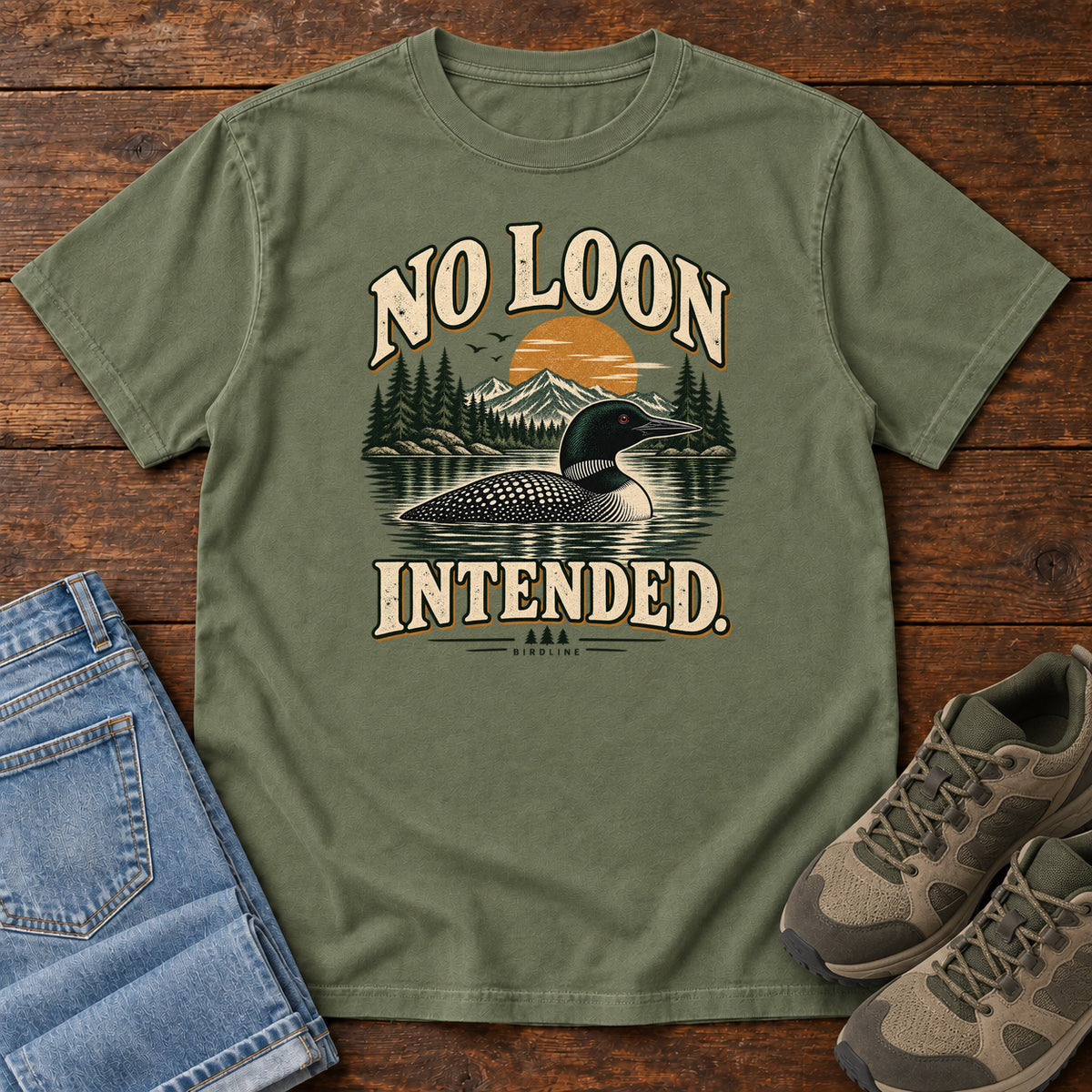 Moss No Loon Intended T-Shirt with a black-and-white waterbird on a mountain lake beneath an orange sun and cream lettering, front flat lay on wood.