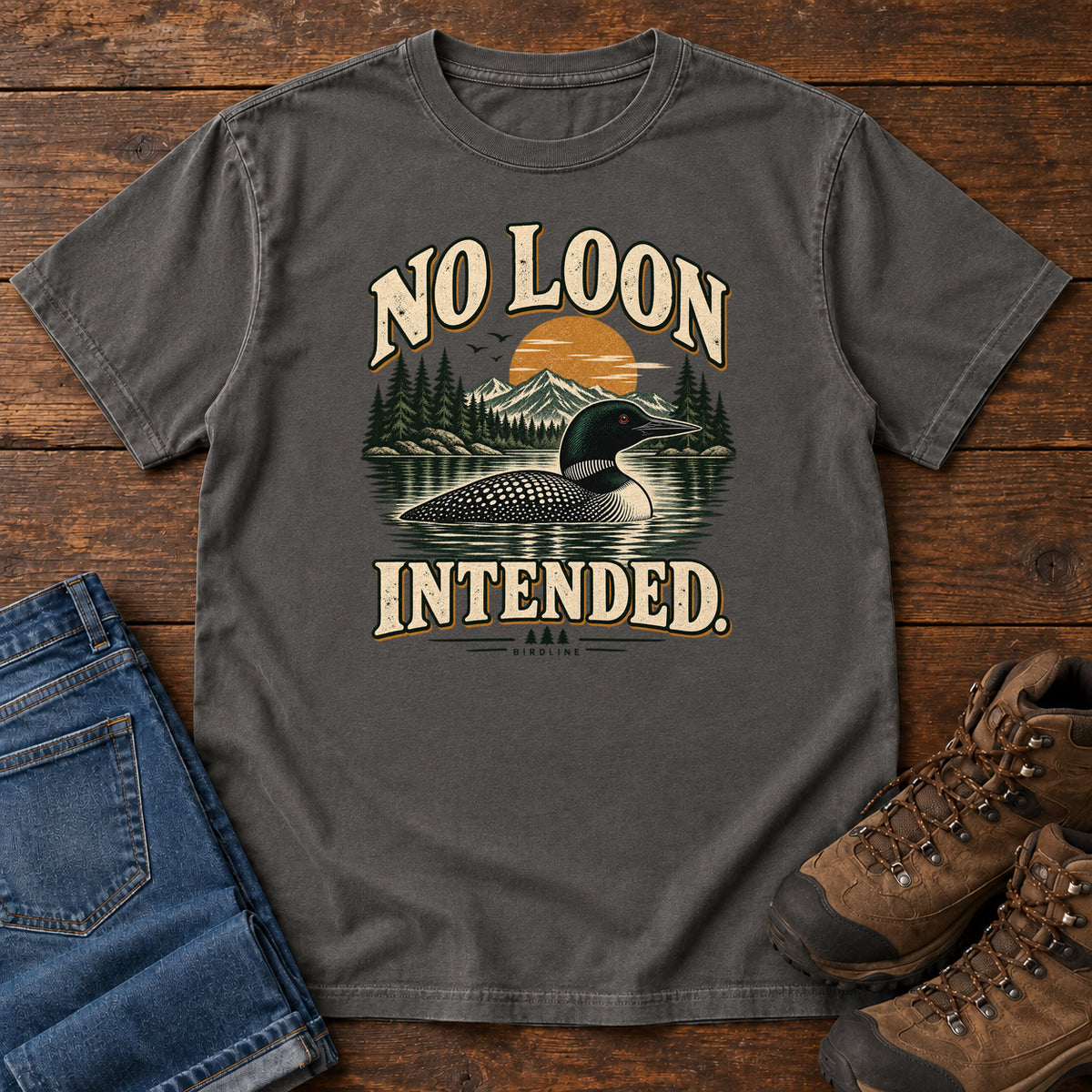 Pepper No Loon Intended T-Shirt with a black-and-white waterbird on a mountain lake beneath an orange sun and cream lettering, front flat lay on wood.