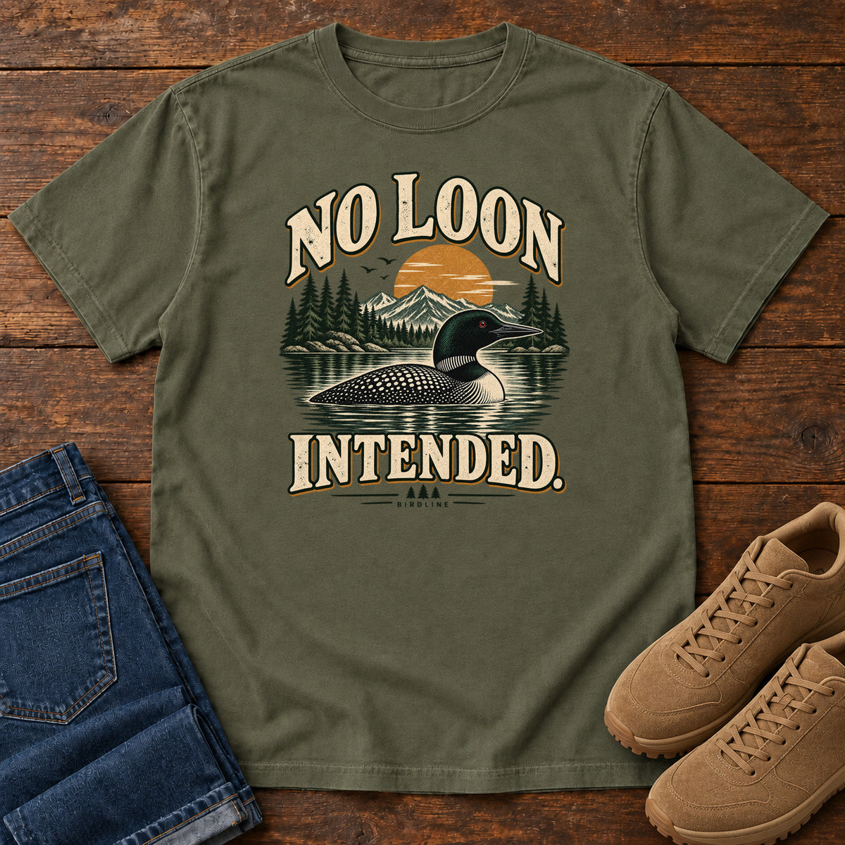 Sage No Loon Intended T-Shirt with a black-and-white waterbird on a mountain lake beneath an orange sun and cream lettering, front flat lay on wood.