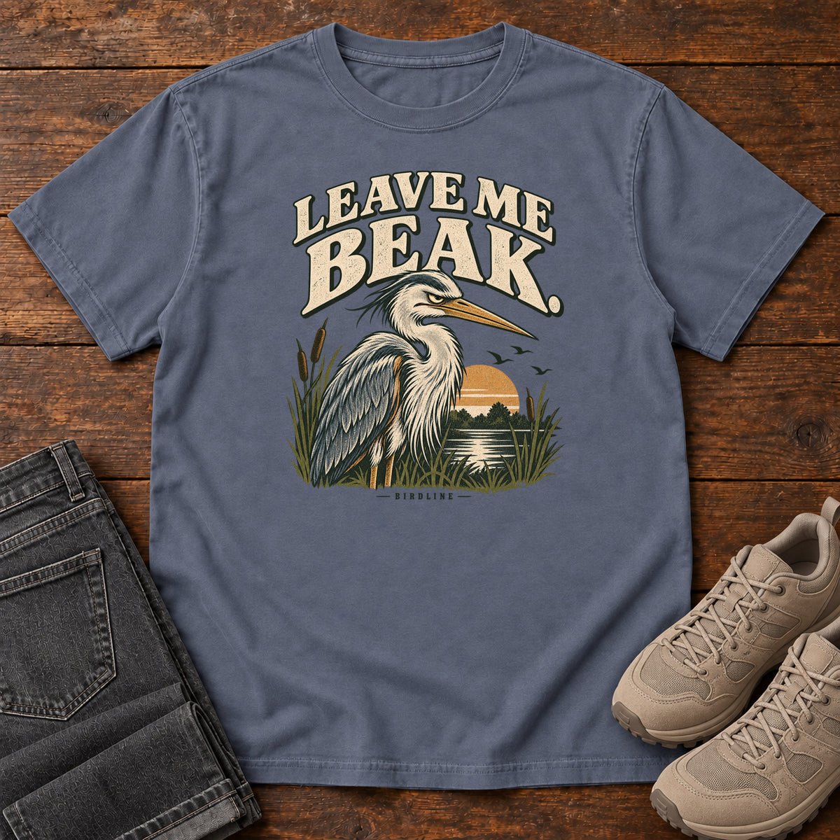 Leave Me Beak T-Shirt in Denim, front laid flat on wood, featuring a gray, long-billed wading bird among reeds beside a sunset, beneath cream lettering.
