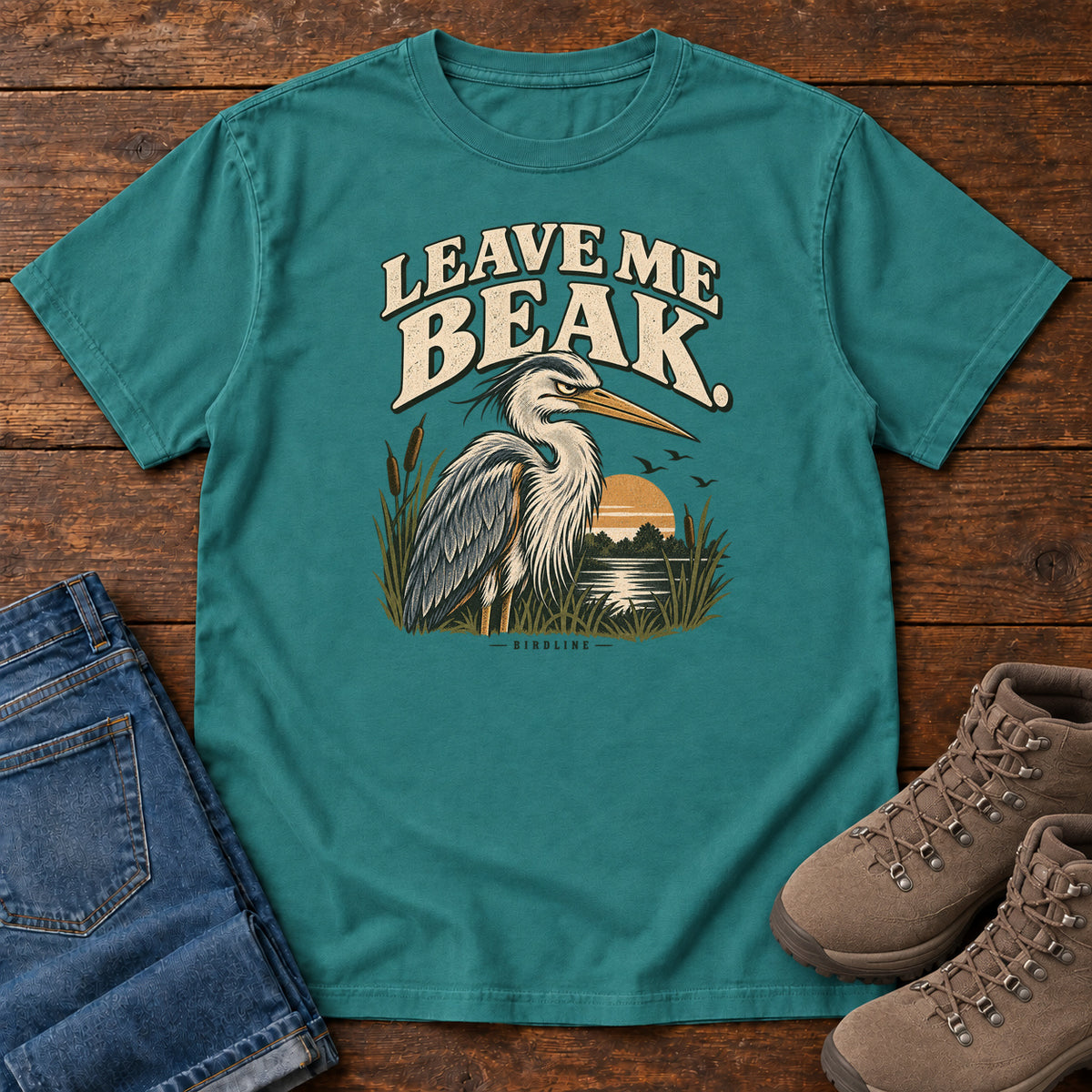 Leave Me Beak T-Shirt in Emerald, front laid flat on wood, featuring a gray, long-billed wading bird among reeds beside a sunset, beneath cream lettering.