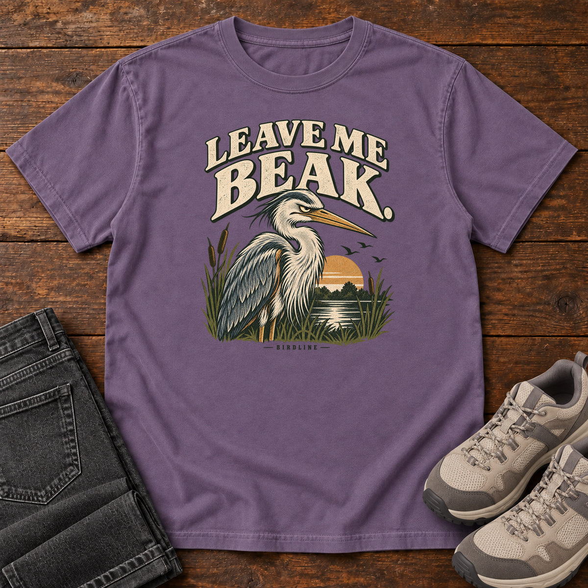 Leave Me Beak T-Shirt in Grape, front laid flat on wood, featuring a gray, long-billed wading bird among reeds beside a sunset, beneath cream lettering.