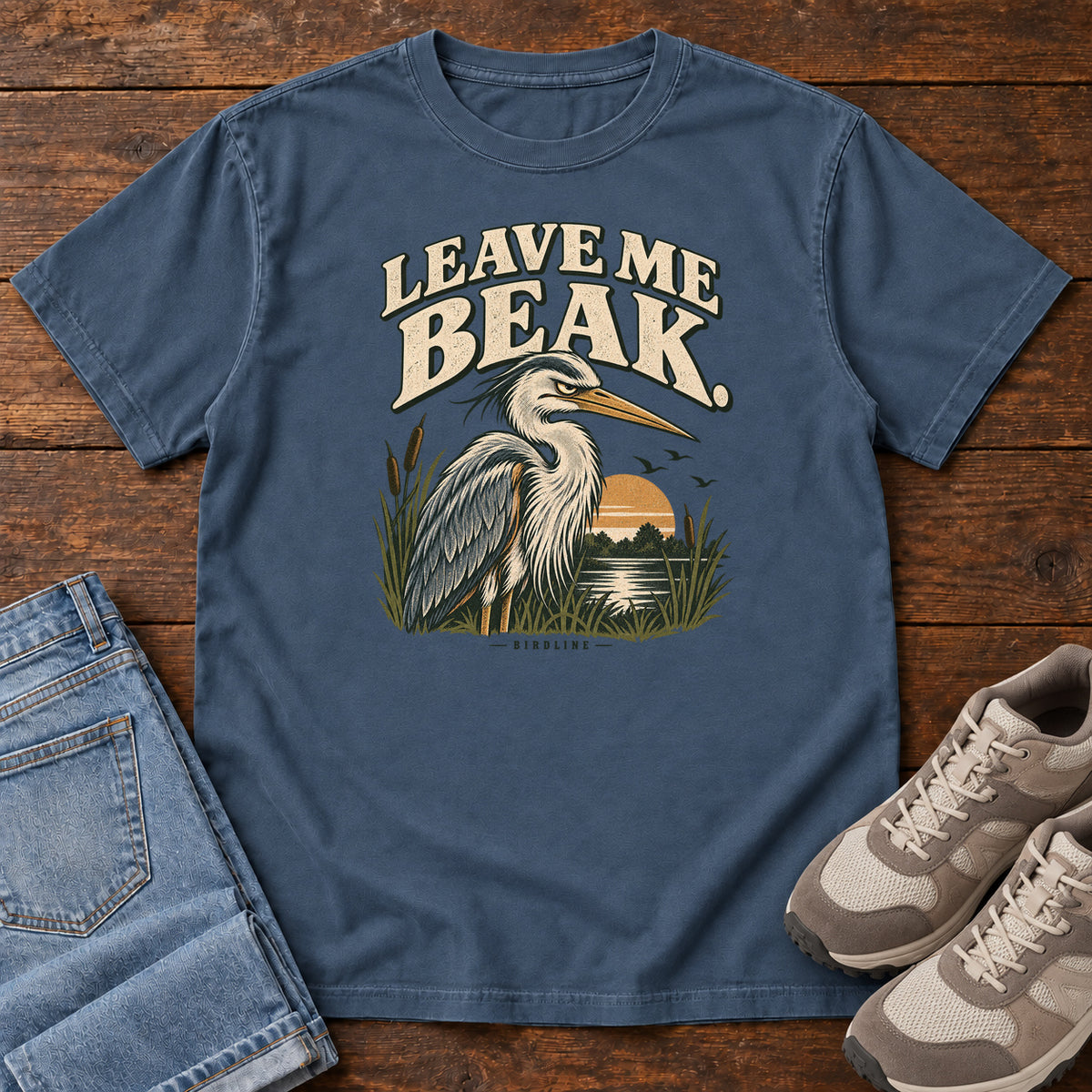 Leave Me Beak T-Shirt in Midnight, front laid flat on wood, featuring a gray, long-billed wading bird among reeds beside a sunset, beneath cream lettering.
