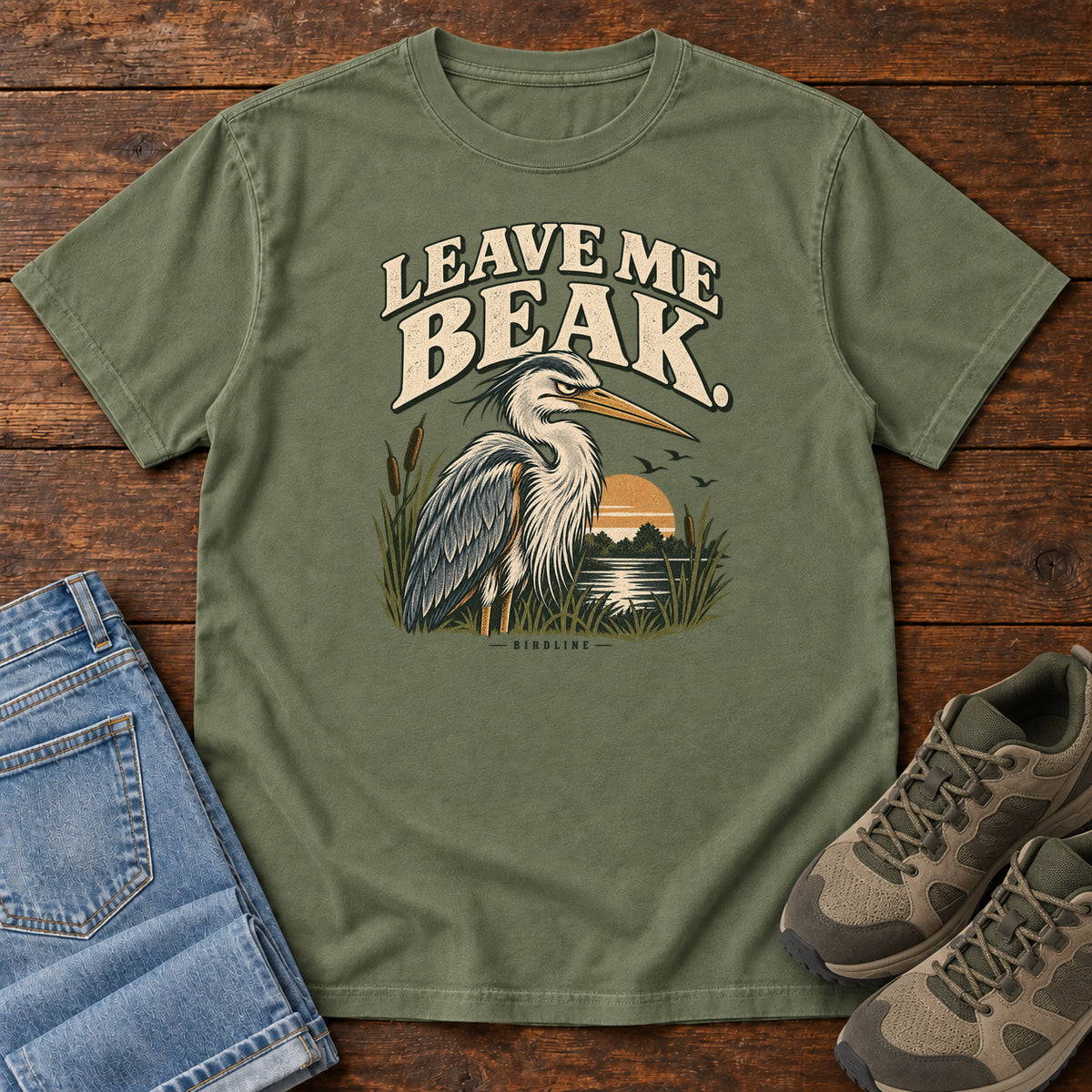 Leave Me Beak T-Shirt in Moss, front laid flat on wood, featuring a gray, long-billed wading bird among reeds beside a sunset, beneath cream lettering.