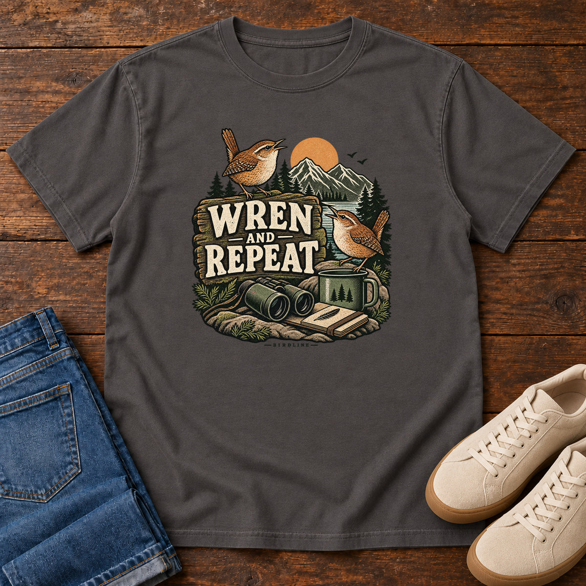 Graphite Wren and Repeat T-Shirt with two small brown birds, binoculars and a camp mug around Wren and Repeat lettering, front flat lay on wood.