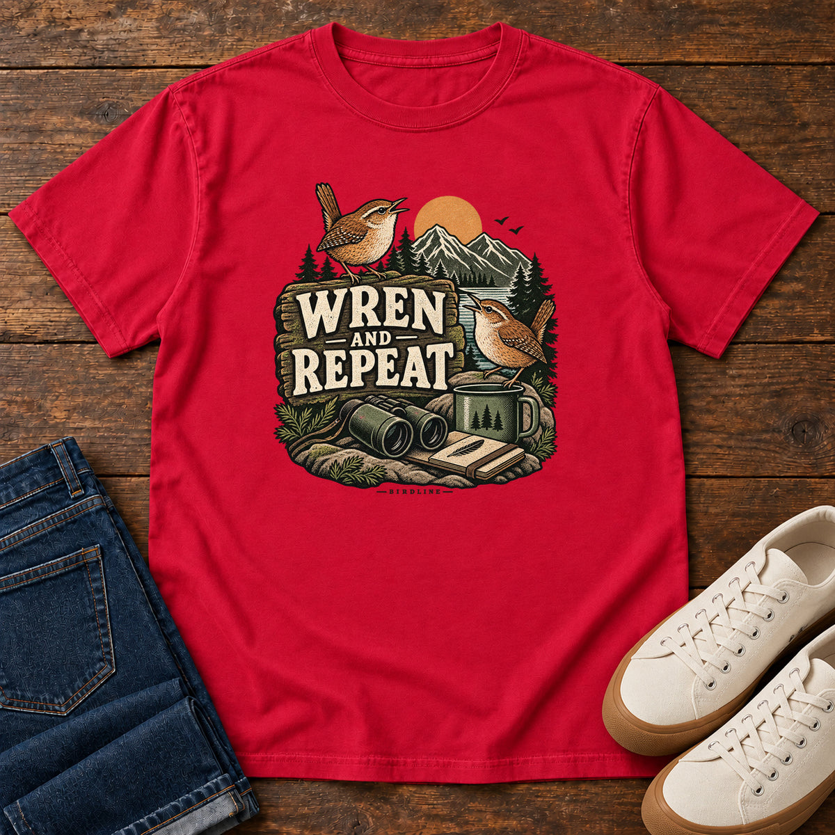 Red Wren and Repeat T-Shirt with two small brown birds, binoculars and a camp mug around Wren and Repeat lettering, front flat lay on wood.