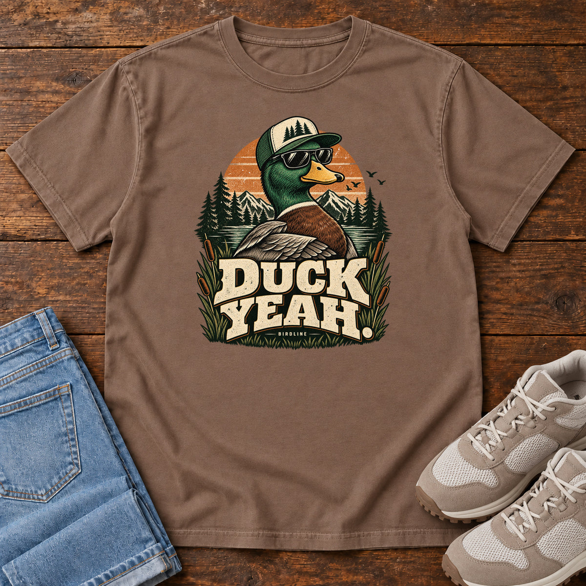 Espresso Duck Yeah T-Shirt with a green-headed duck wearing a cap and sunglasses above bold cream lettering, front flat lay on wood.