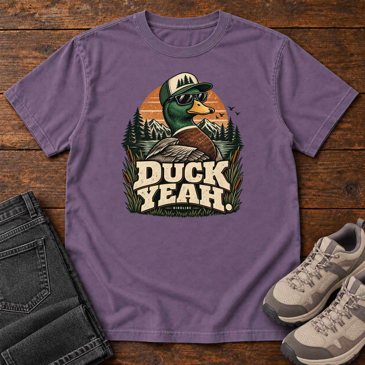 Grape Duck Yeah T-Shirt with a green-headed duck wearing a cap and sunglasses above bold cream lettering, front flat lay on wood.