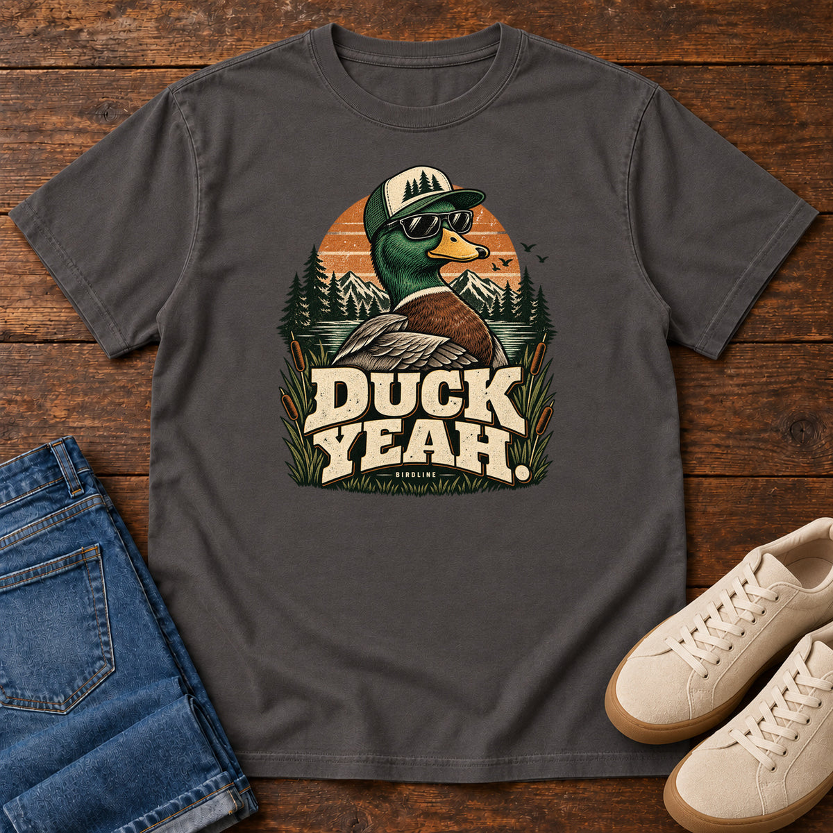 Graphite Duck Yeah T-Shirt with a green-headed duck wearing a cap and sunglasses above bold cream lettering, front flat lay on wood.