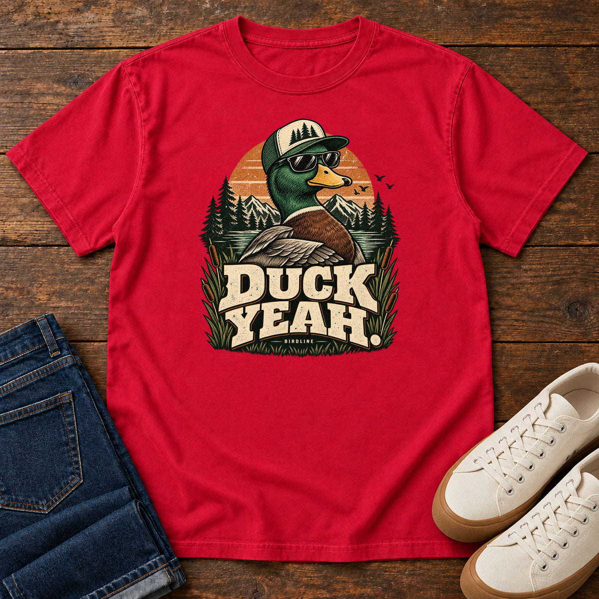 Red Duck Yeah T-Shirt with a green-headed duck wearing a cap and sunglasses above bold cream lettering, front flat lay on wood.