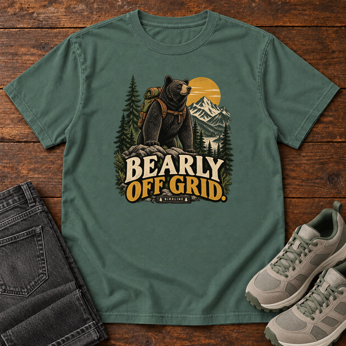 Blue Spruce Bearly Off Grid T-Shirt with a black bear wearing a backpack before mountains, pines and a golden sun; front flat lay on wood.
