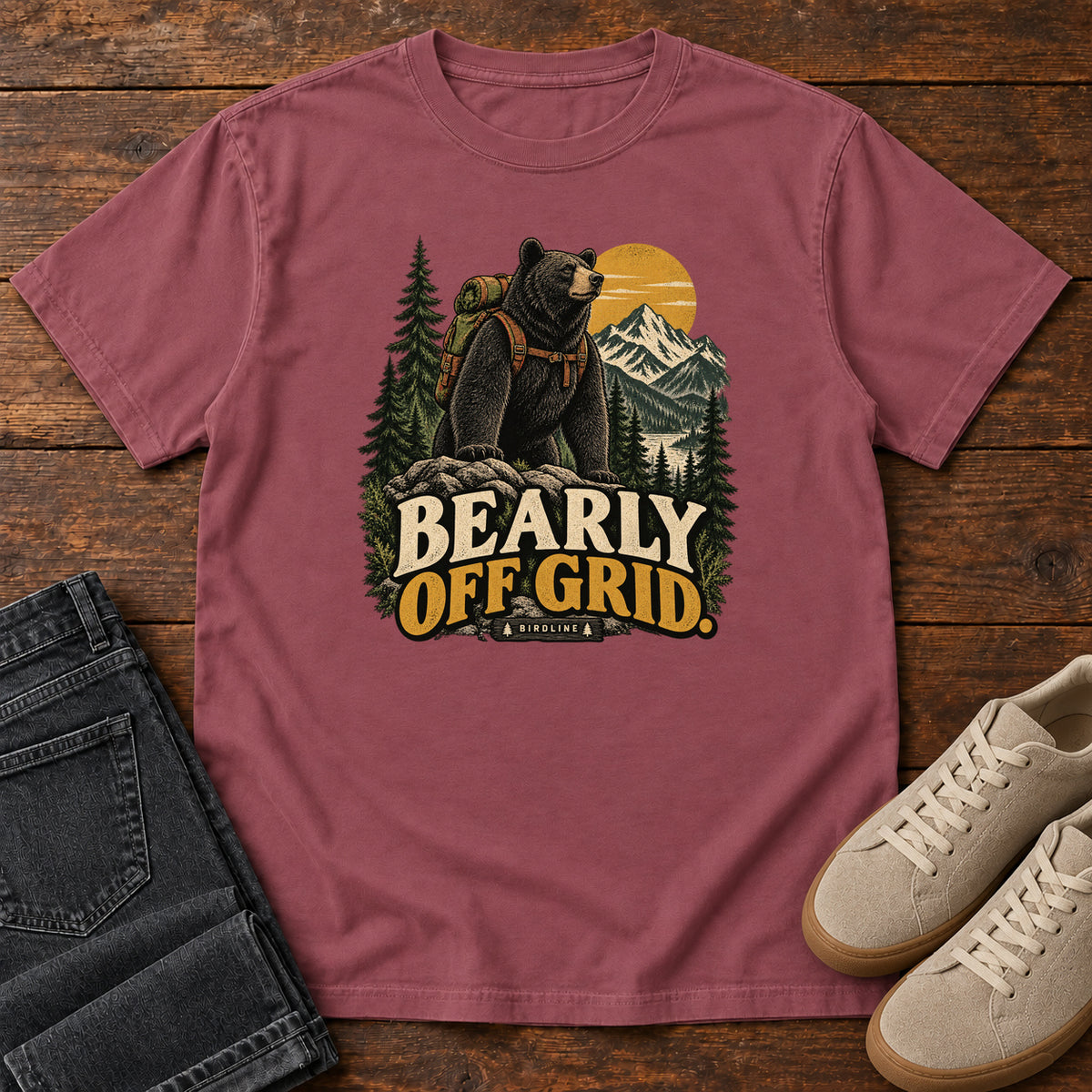 Brick Bearly Off Grid T-Shirt with a black bear wearing a backpack before mountains, pines and a golden sun; front flat lay on wood.