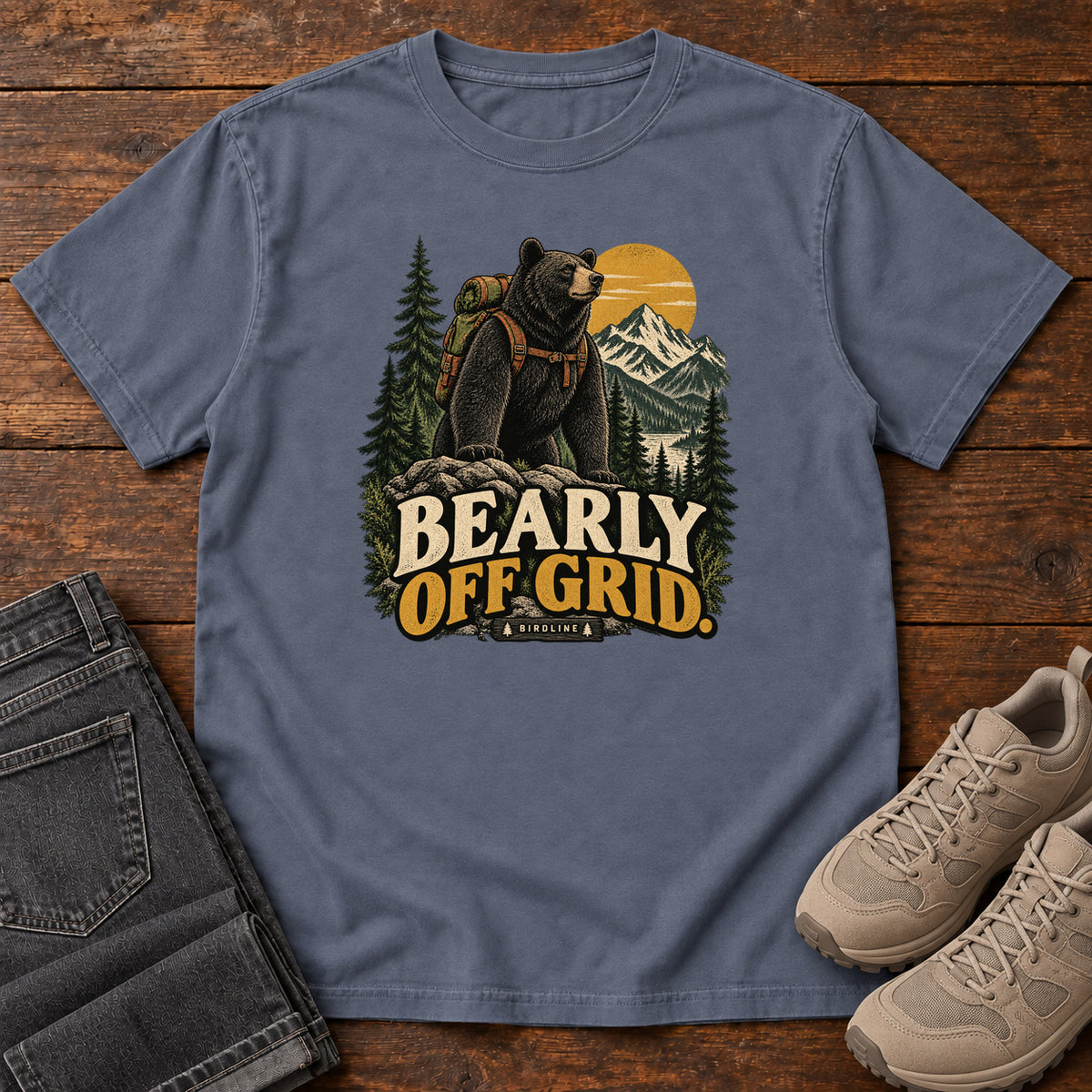 Denim Bearly Off Grid T-Shirt with a black bear wearing a backpack before mountains, pines and a golden sun; front flat lay on wood.