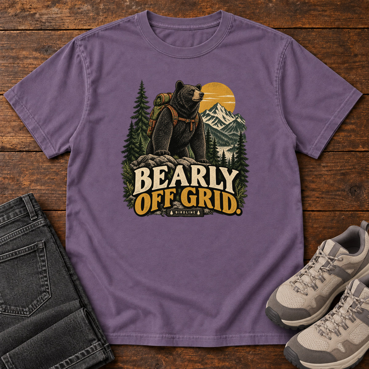 Grape Bearly Off Grid T-Shirt with a black bear wearing a backpack before mountains, pines and a golden sun; front flat lay on wood.