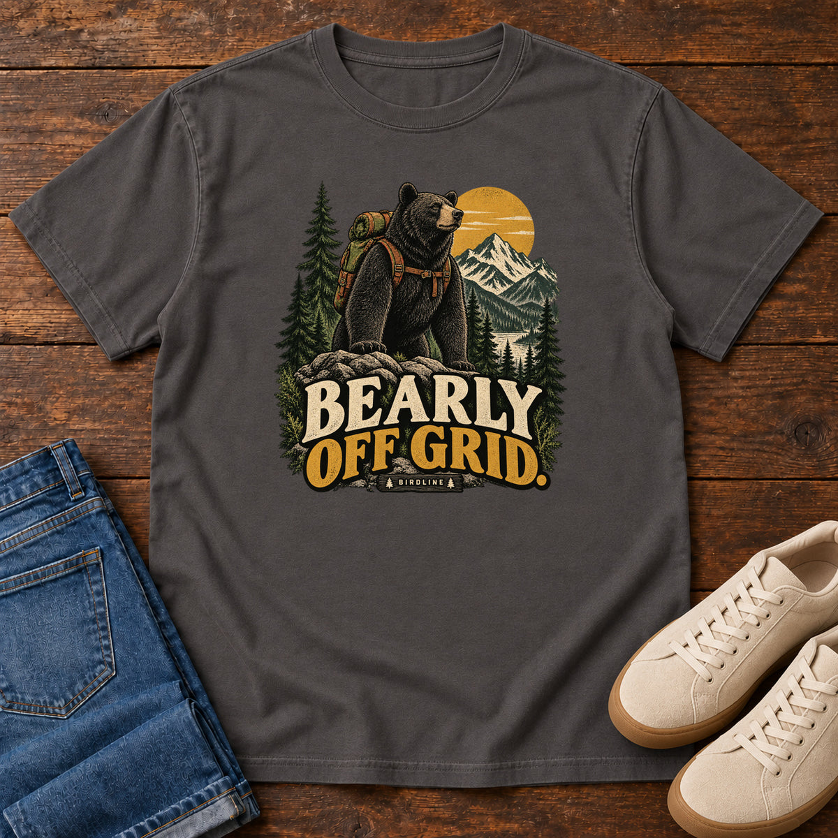 Graphite Bearly Off Grid T-Shirt with a black bear wearing a backpack before mountains, pines and a golden sun; front flat lay on wood.