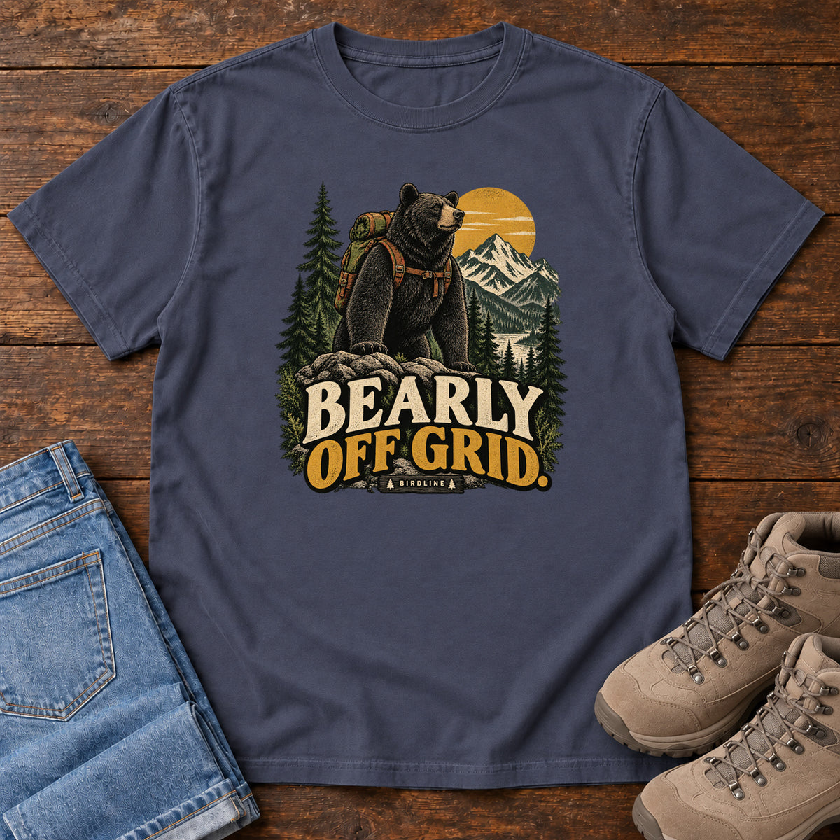 Navy Bearly Off Grid T-Shirt with a black bear wearing a backpack before mountains, pines and a golden sun; front flat lay on wood.