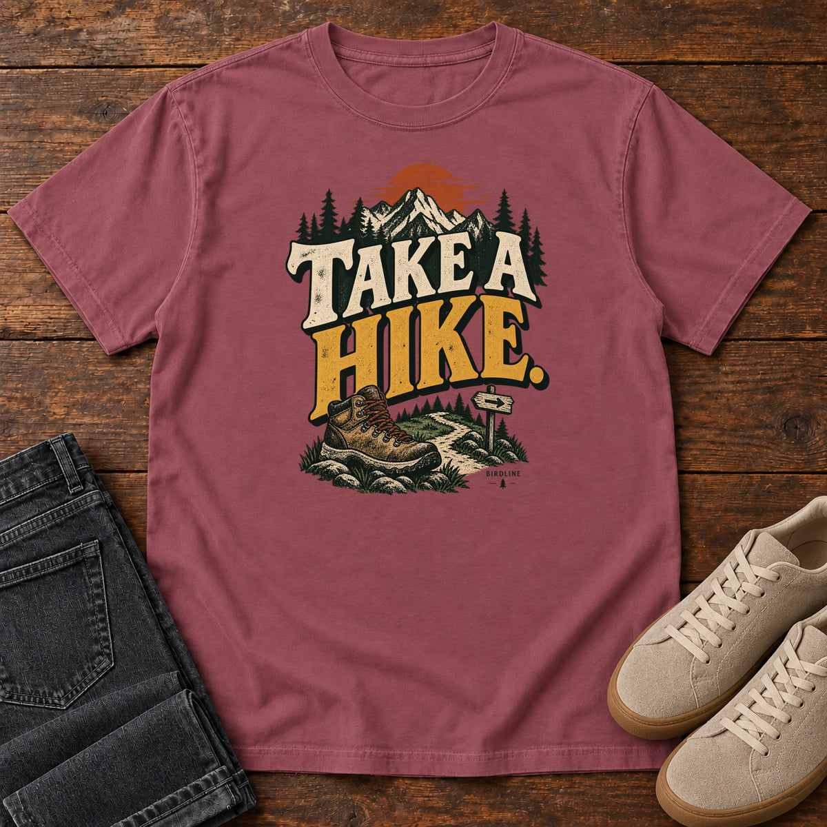 Brick Take a Hike T-Shirt with a hiking boot, mountain trail and orange sun around bold cream and gold lettering; front flat lay on wood.